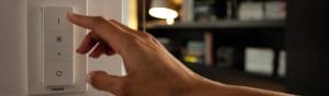 „Hand, die den intelligenten LED-Dimmschalter von Philips einstellt, der die Raumbeleuchtung in einem gemütlichen Wohnzimmer mit Bücherregalen steuert.“
