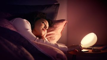 Eine Frau, die im Bett liegt, greift zum Nachttisch, wo eine Tischlampe neben ihrem Telefon steht